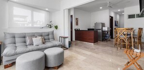 Una sala de estar moderna con un sofá gris, otomanas, un comedor con sillas de madera, una cocina con refrigerador y un televisor montado en la pared.