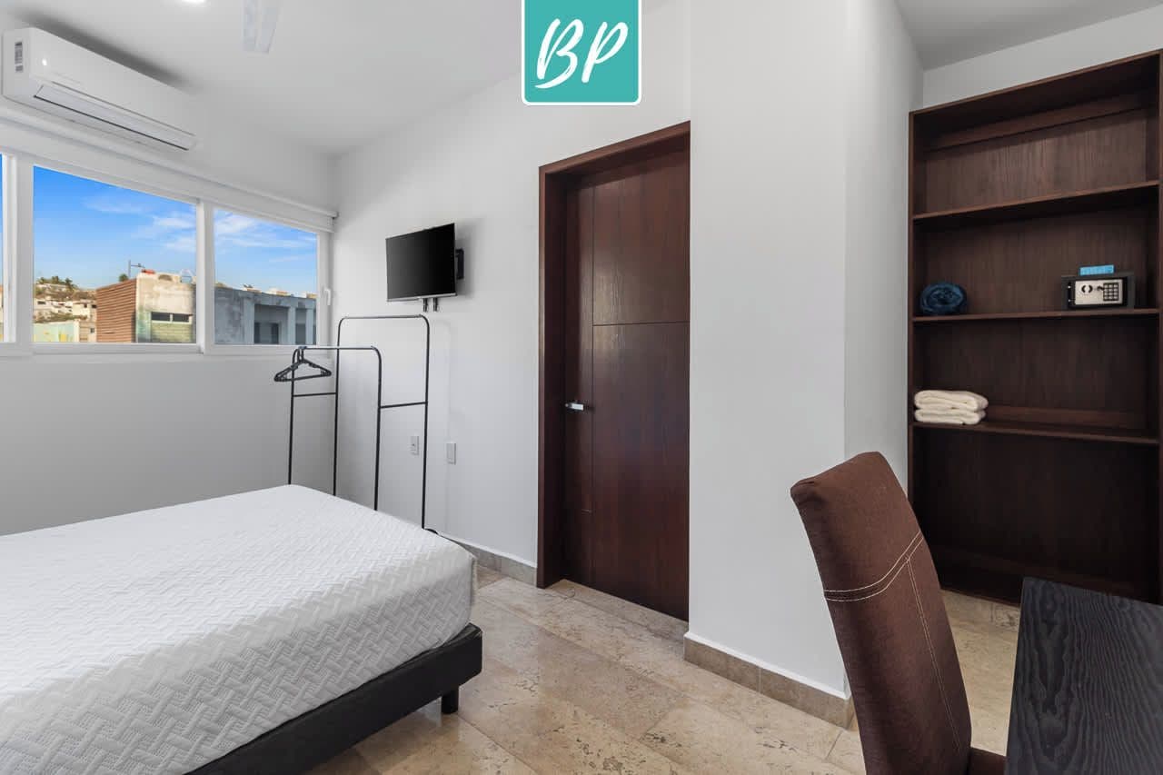 Un dormitorio con cama, televisor montado en la pared, armario y ventana con vistas a edificios.