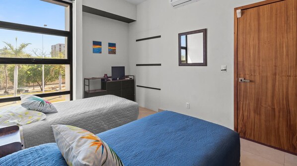 A bedroom with a bed, a desk, a chair, a wardrobe, and a window with a view of trees and buildings.