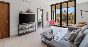 A modern living room with a flat-screen TV, a coffee table, a sofa, and a large window offering a view of the outdoors. - 2BR, Rooftop Pool w/ Views, & Parking: Vista 101 (Bucerías)