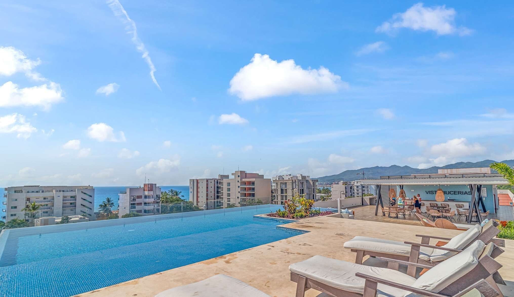 A rooftop pool area with lounge chairs, a restaurant, and a view of the ocean and cityscape.