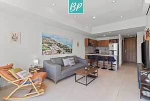 A modern living room with a grey sofa, a coffee table, and a rocking chair. There is a kitchen area with a fridge, microwave, and sink. A large painting of a cityscape is on the wall.
