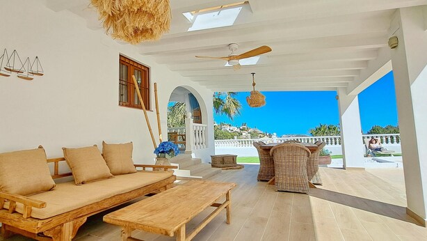 Un salón espacioso con sofá de madera, una mesa de madera y muebles de mimbre. Se ve el exterior a través de un arco abierto.