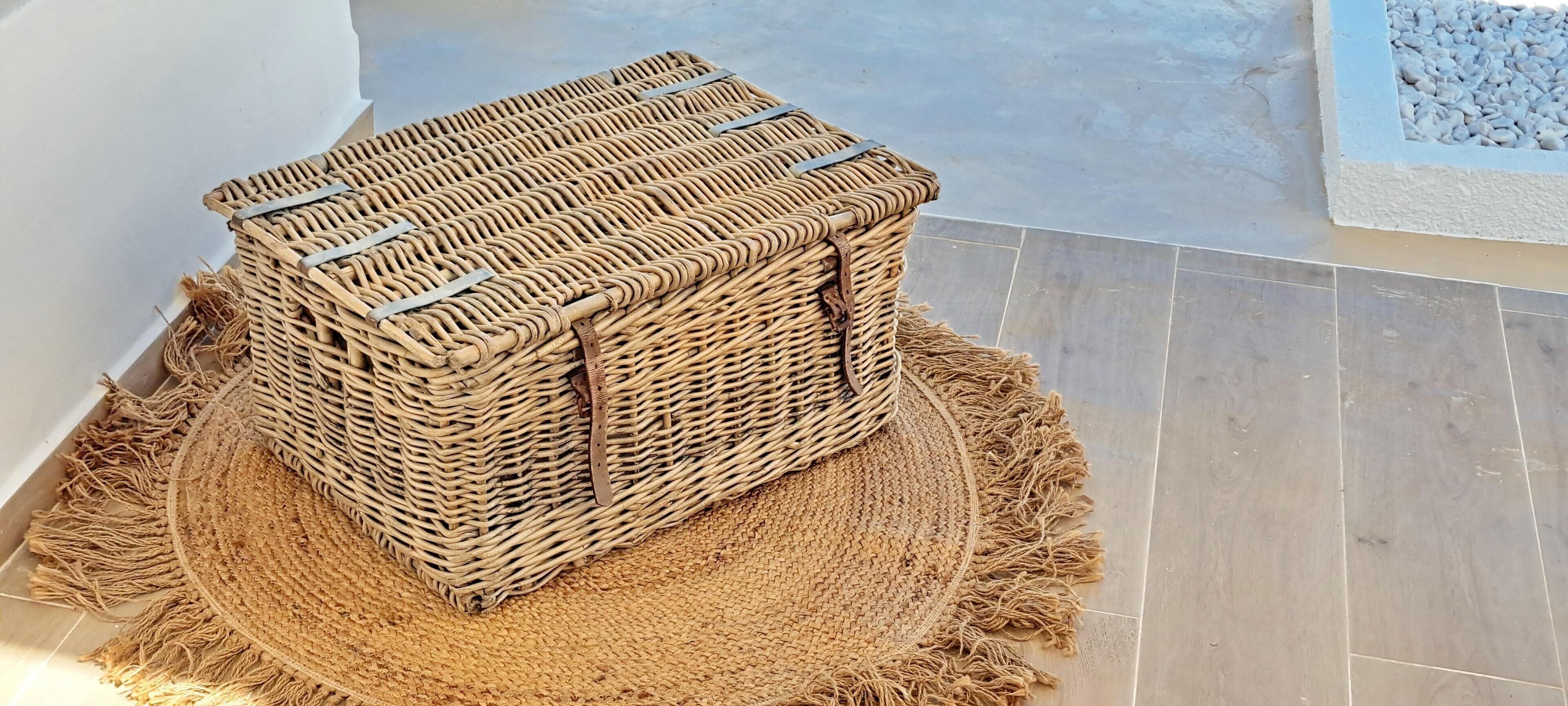 A wicker basket with a handle, placed on a woven mat on a tiled floor.