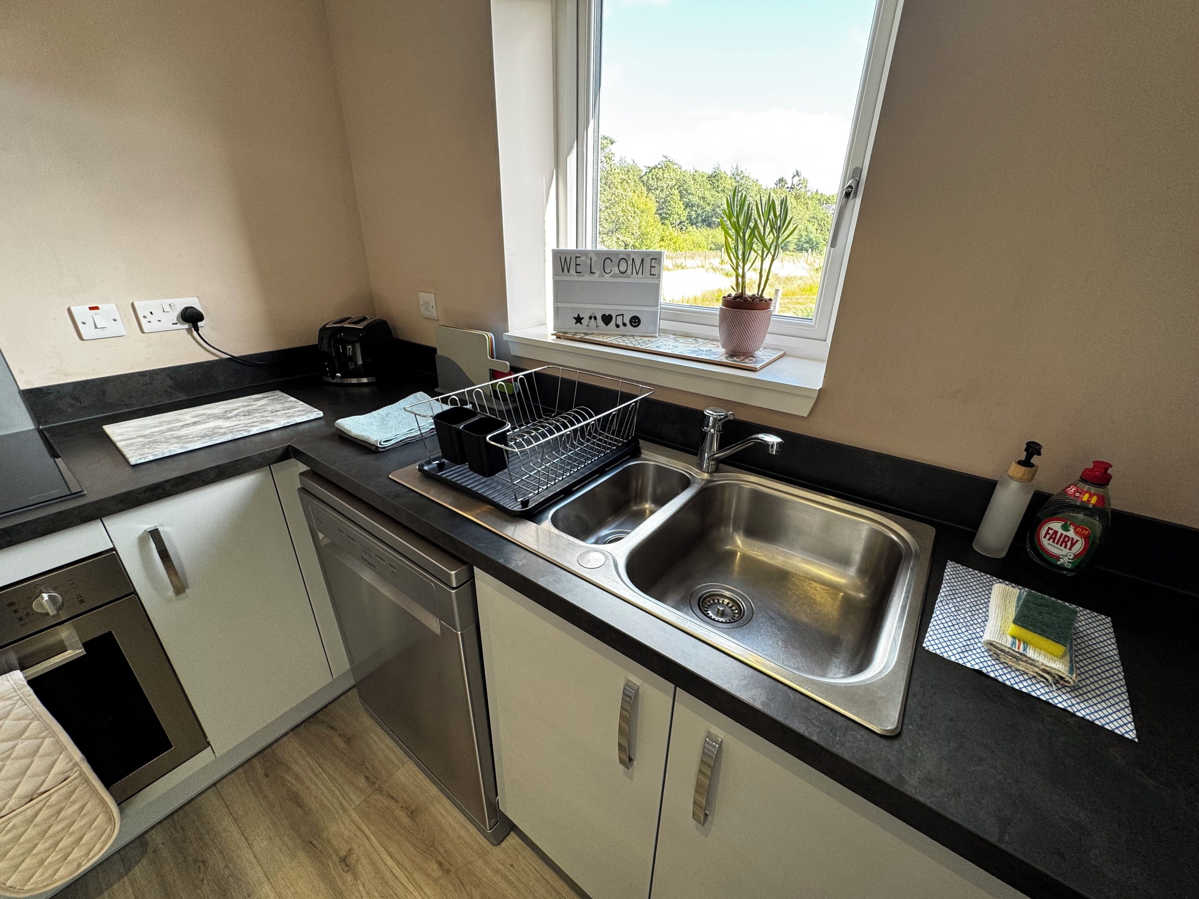 A modern kitchen with stainless steel appliances, a black countertop, and a window with a view of greenery.