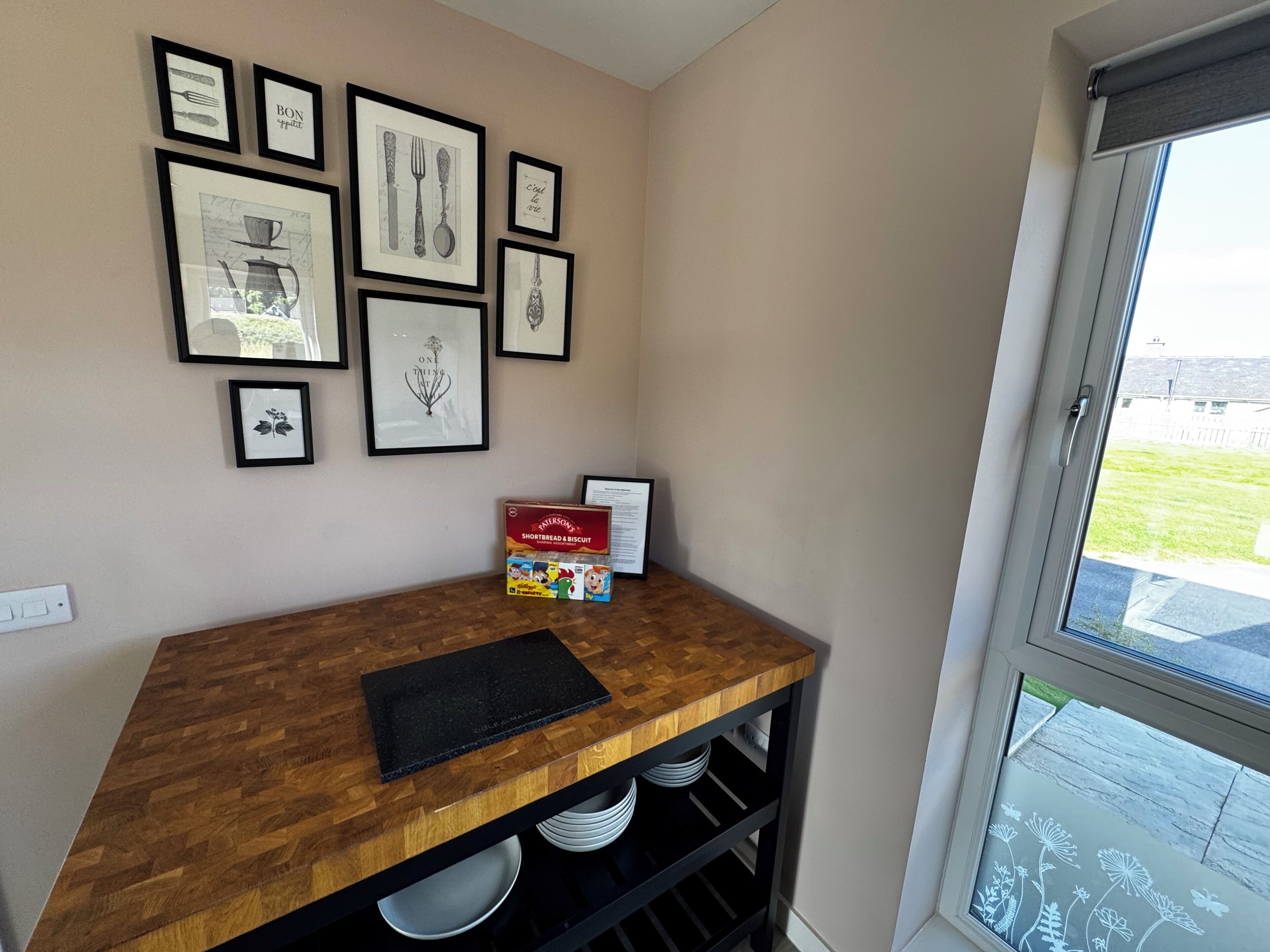 A kitchen with a wooden countertop, framed pictures on the wall, and a window with a view.
