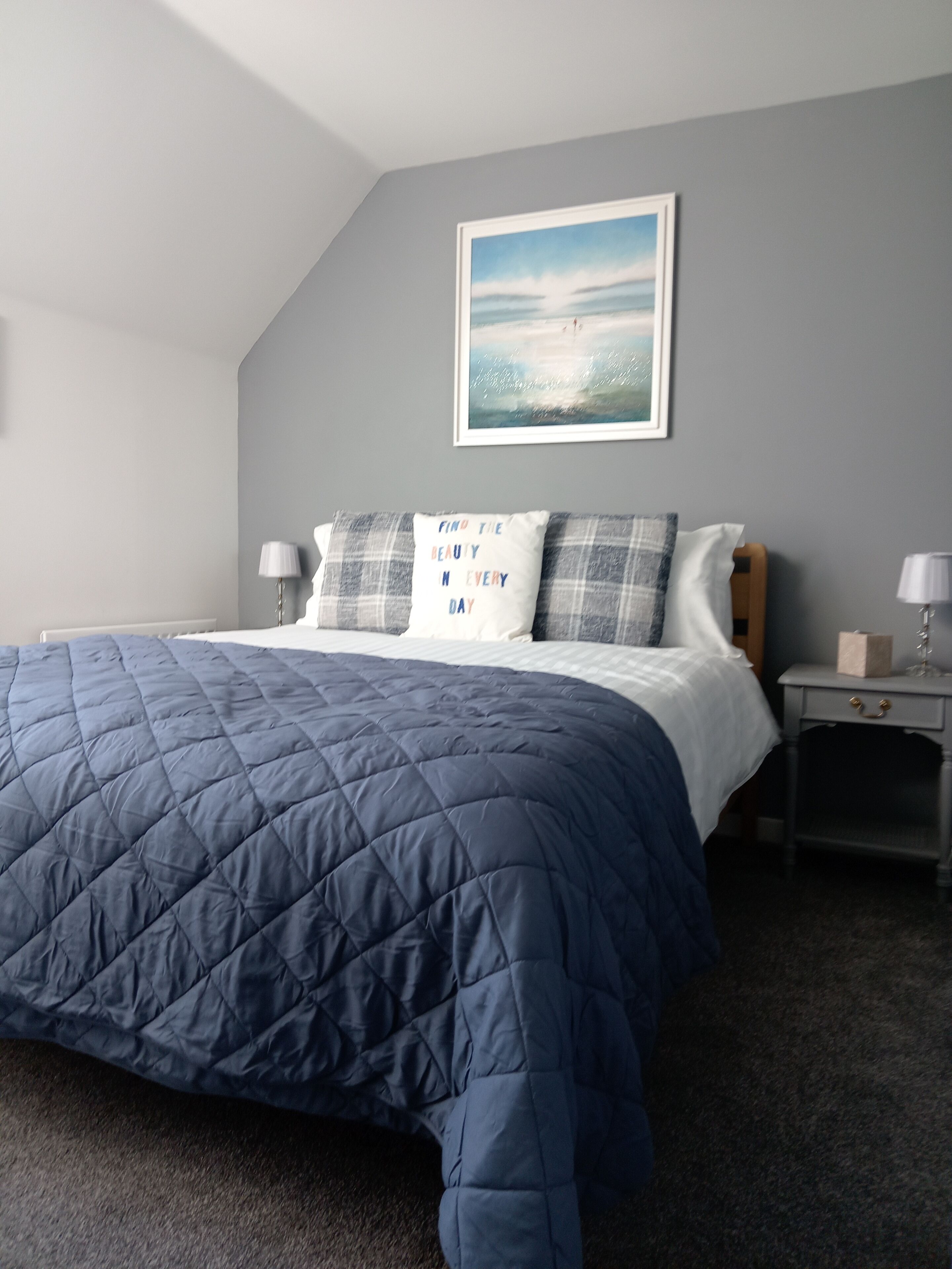 Une chambre à coucher avec un grand lit, des murs gris, un tableau encadré au-dessus du lit et deux tables de chevet avec des lampes.