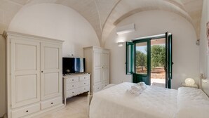 Ein Schlafzimmer mit einem großen Bett, einem Fernseher auf einer Kommode und Blick ins Freie durch eine offene Tür.