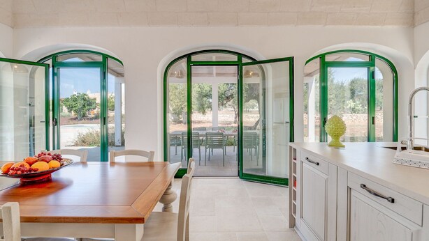 Eine moderne Küche mit Essbereich, ausgestattet mit einem Holztisch, weißen Stühlen und Blick auf die Außenpatio durch grüne Glastüren.