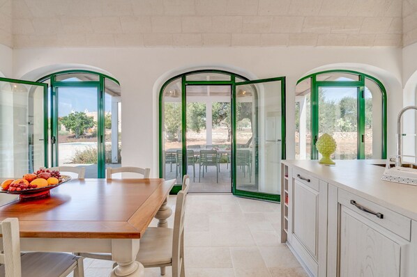 A modern kitchen with a dining area, featuring a wooden table, white chairs, and a view of the outdoor patio through green-framed glass doors.