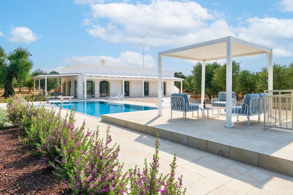 Zona junto a la piscina con un área de estar cubierta, una piscina y un edificio con ventanas arqueadas.