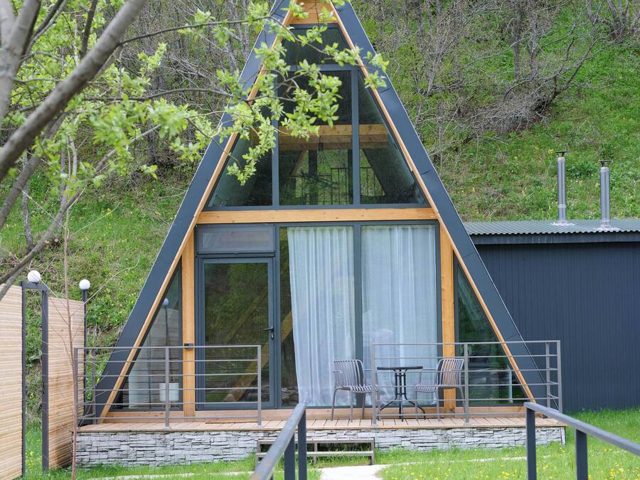 A modern A-frame cabin with a glass front, wooden deck, and a stone wall in front.