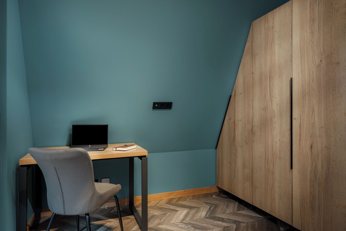 A room with teal walls, a wooden desk with a laptop, a grey chair, and a herringbone-patterned floor.