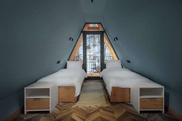 Un dormitorio con dos camas, suelo de madera con patrón de espiga y vistas a un paisaje nevado a través de un ventanal triangular.