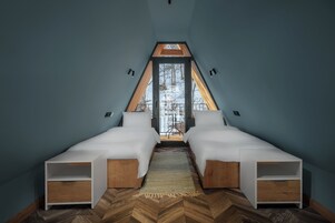 Un dormitorio con dos camas, suelo de madera con patrón de espiga y vistas a un paisaje nevado a través de un ventanal triangular.