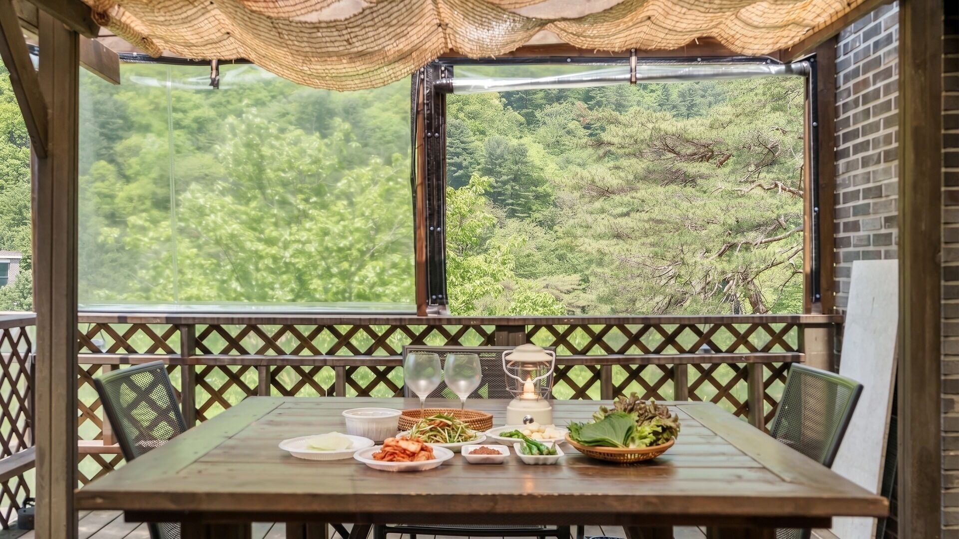 A wooden table with food and drinks, a lantern, and a view of a forest through a window.