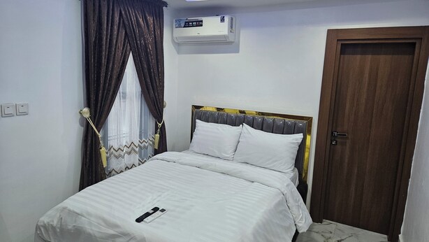 A neatly made bed with white bedding, a remote control, and a wall-mounted air conditioner. - Bimsy's Place  (Abuja)