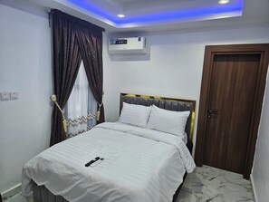 A neatly made bed with white bedding, a remote control, and a wall-mounted air conditioner. - Bimsy's Place  (Abuja)