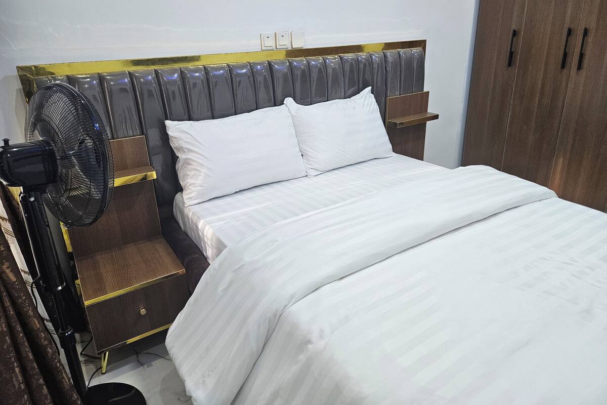 Un letto rifatto con biancheria bianca, una testata scura e una ventola sul comodino.