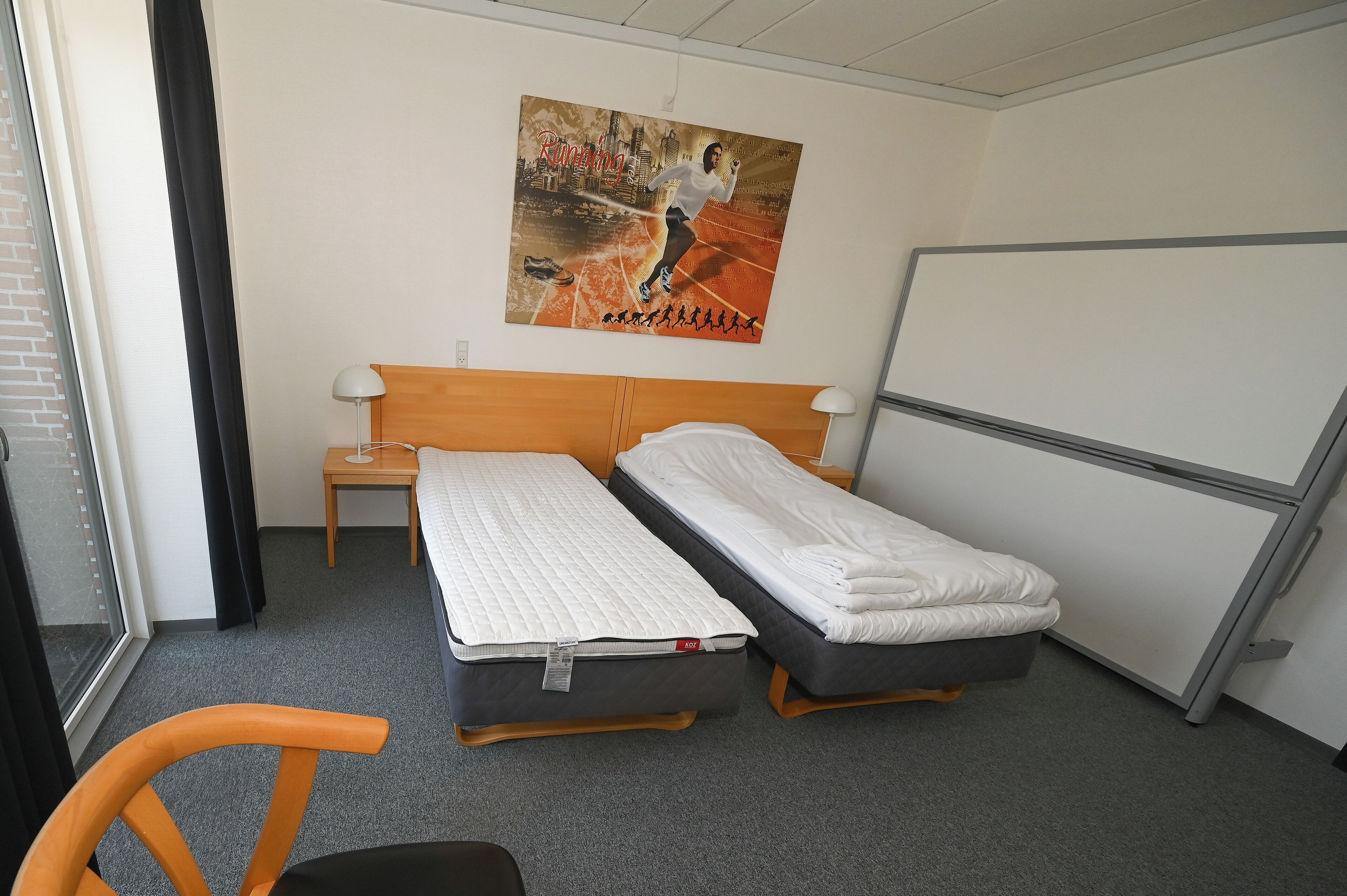 A hotel room with two beds, a nightstand, a lamp, a window with curtains, and a poster on the wall.