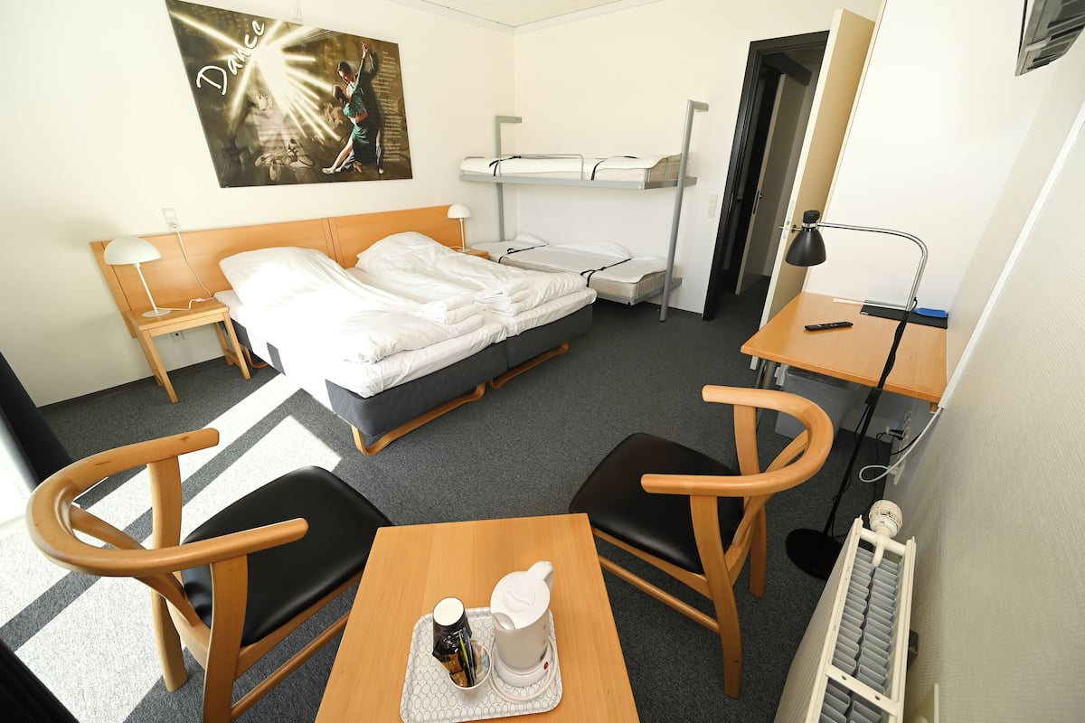 A hotel room with a bed, two bedside tables, a desk, two chairs, a radiator, and a wall-mounted artwork.