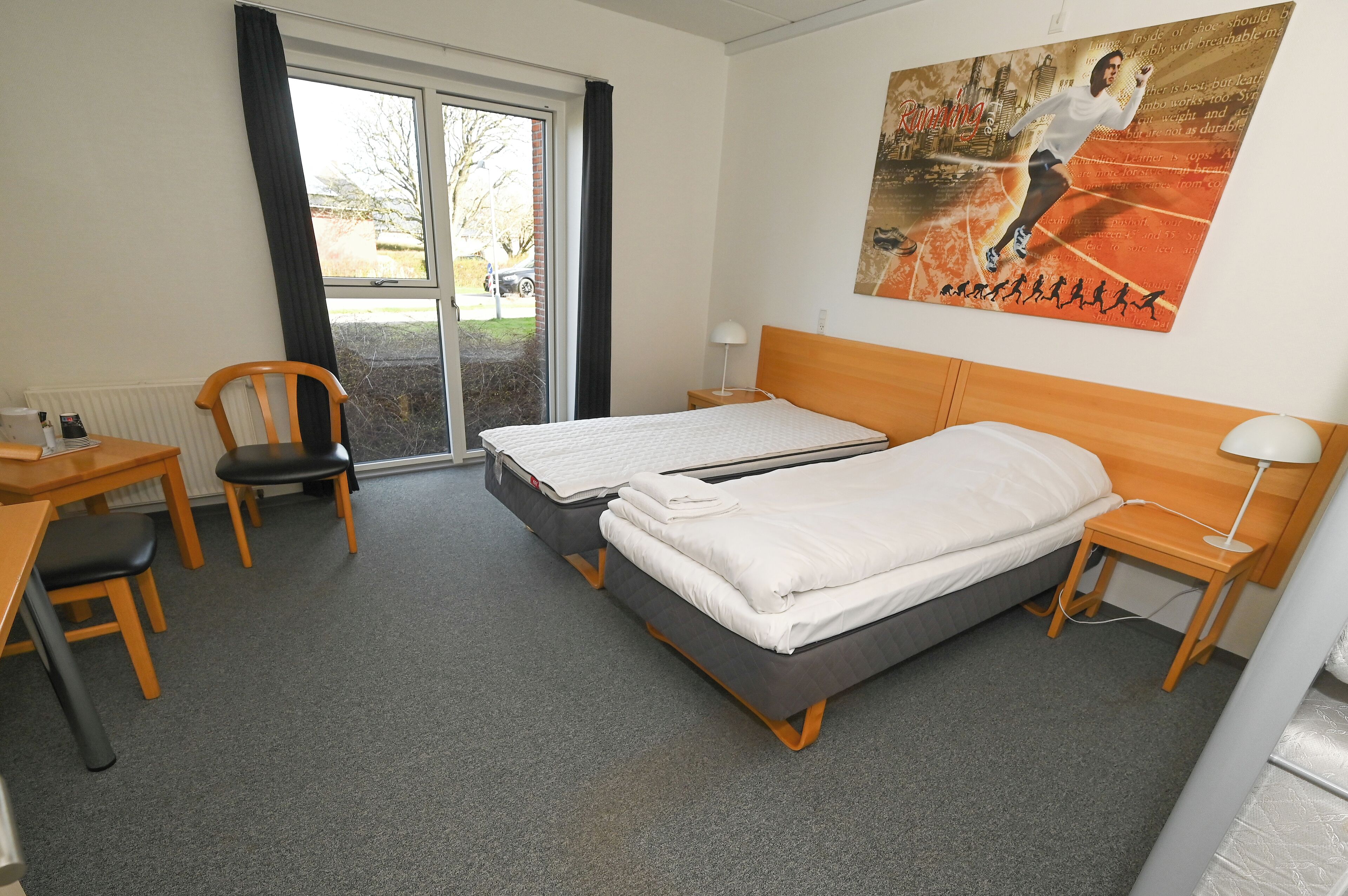 Ein Hotelzimmer mit zwei Einzelbetten, einem Holztisch mit Stuhl, einem kleinen Tisch mit Lampe und einem großen Fenster mit Ausblick.