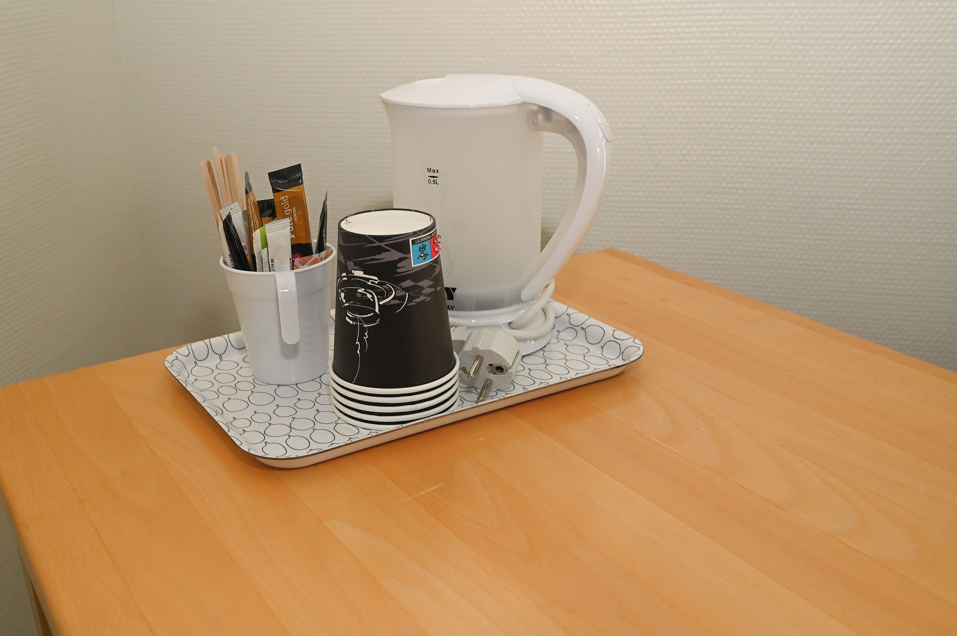 Una cafetera blanca sobre una bandeja, con un vaso de bolígrafos y una taza negra sobre una mesa de madera.