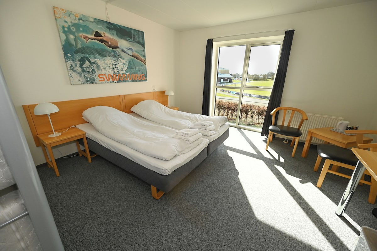 A hotel room with a bed, a desk, a chair, a window with a view of a field, and a painting on the wall.