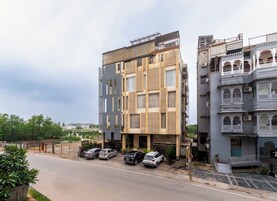 Un edificio de varios pisos con fachada moderna, un estacionamiento con varios coches y un edificio tradicional con balcones ornamentados.