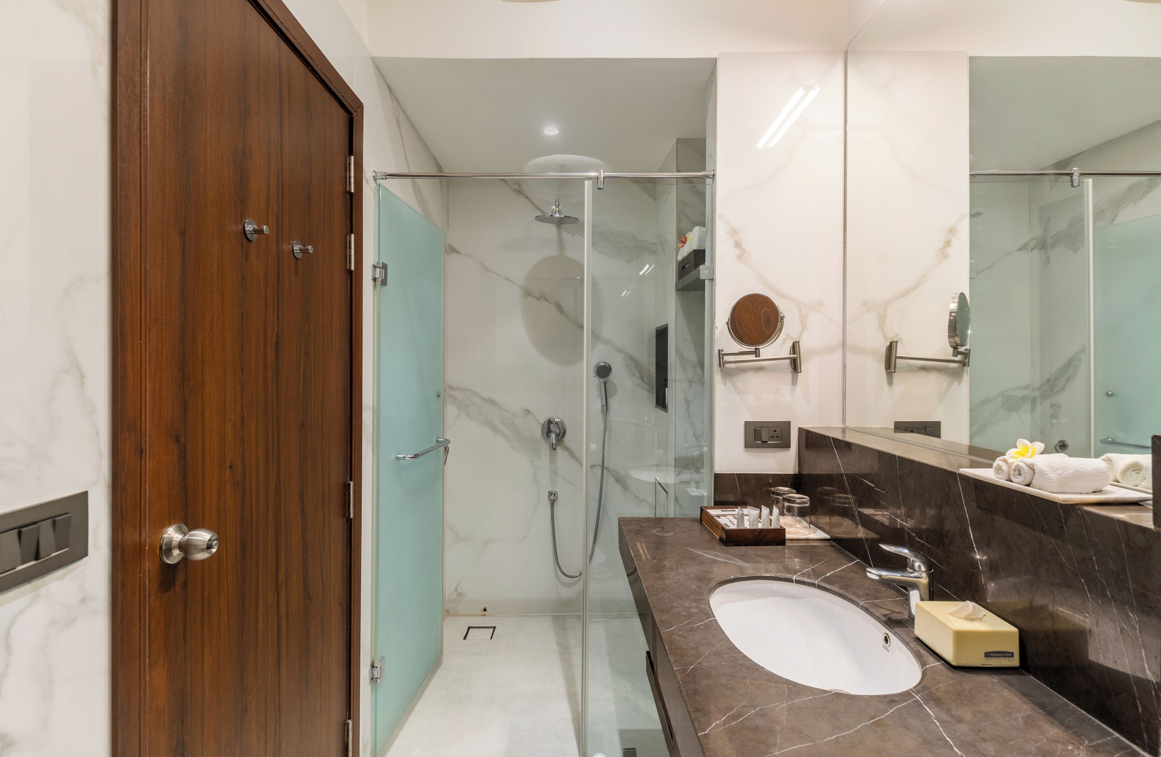 Un baño moderno con puerta de madera, cabina de ducha de vidrio, lavabo de mármol y tocador con artículos de aseo.