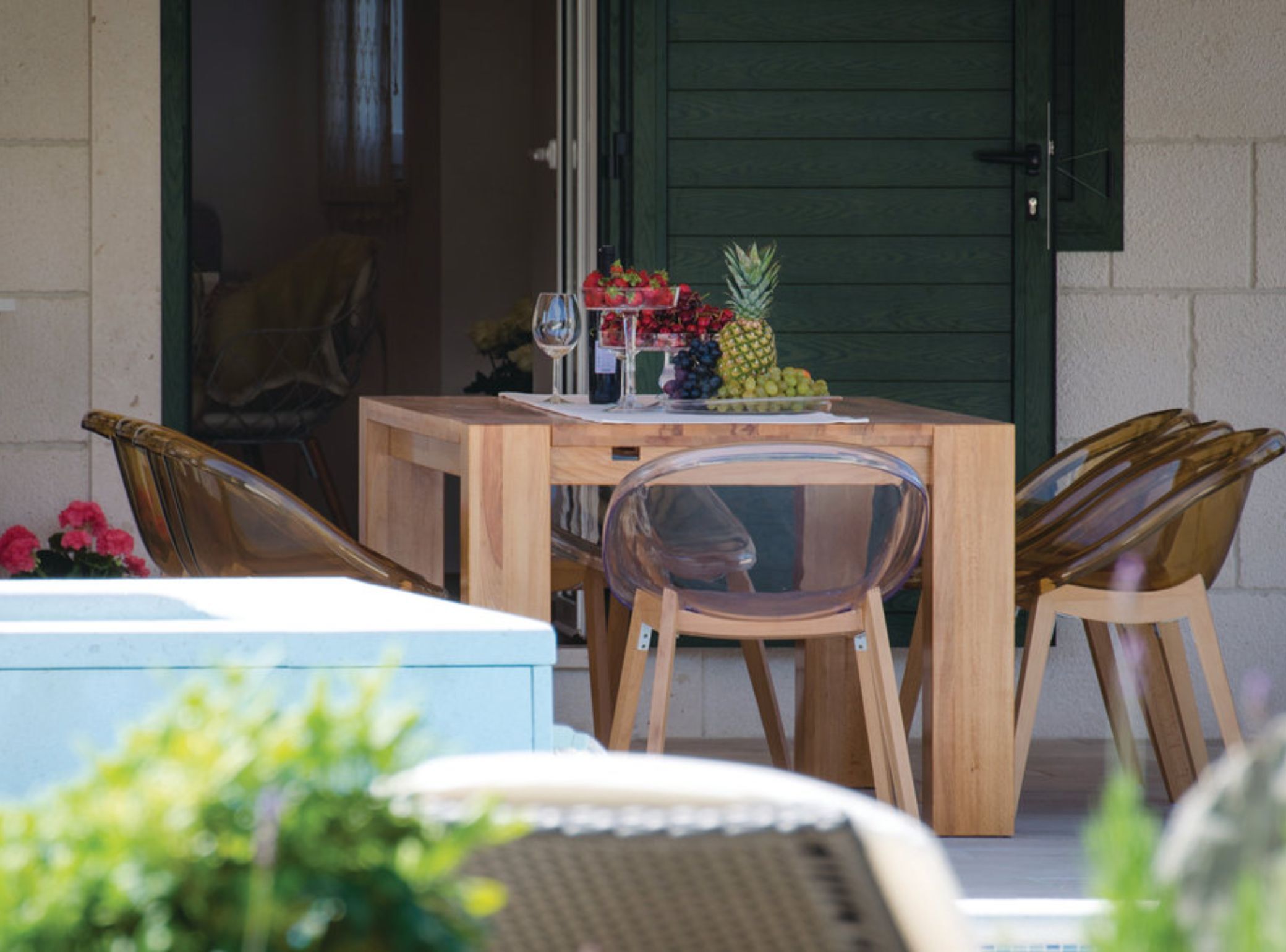 A wooden table with a fruit bowl, wine glasses, and a pineapple. There are two wooden chairs and a clear chair.