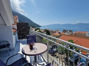 Un balcon avec une table et des chaises, donnant sur une ville côtière aux bâtiments aux toits rouges, et offrant une vue sur la mer et les montagnes.