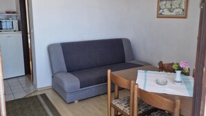 A compact living space with a grey sofa, a wooden dining table with chairs, a small kitchen area, and a framed picture on the wall. - Lora Apartments Gradac (Gradac)