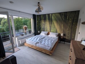 A modern bedroom with a large bed, a view of a forest through a window, and a balcony.