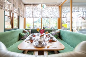 Un'area pranzo con un divano verde, un tavolo in legno apparecchiato per un pasto e vista su un paesaggio verde attraverso ampie finestre.