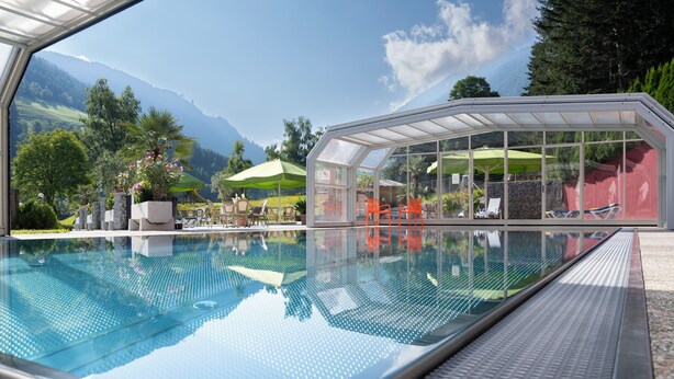 A clear view of a swimming pool with a glass enclosure, surrounded by outdoor seating and a scenic mountain backdrop. - DAS Hubertus Apartments in Schladming (Schladming)