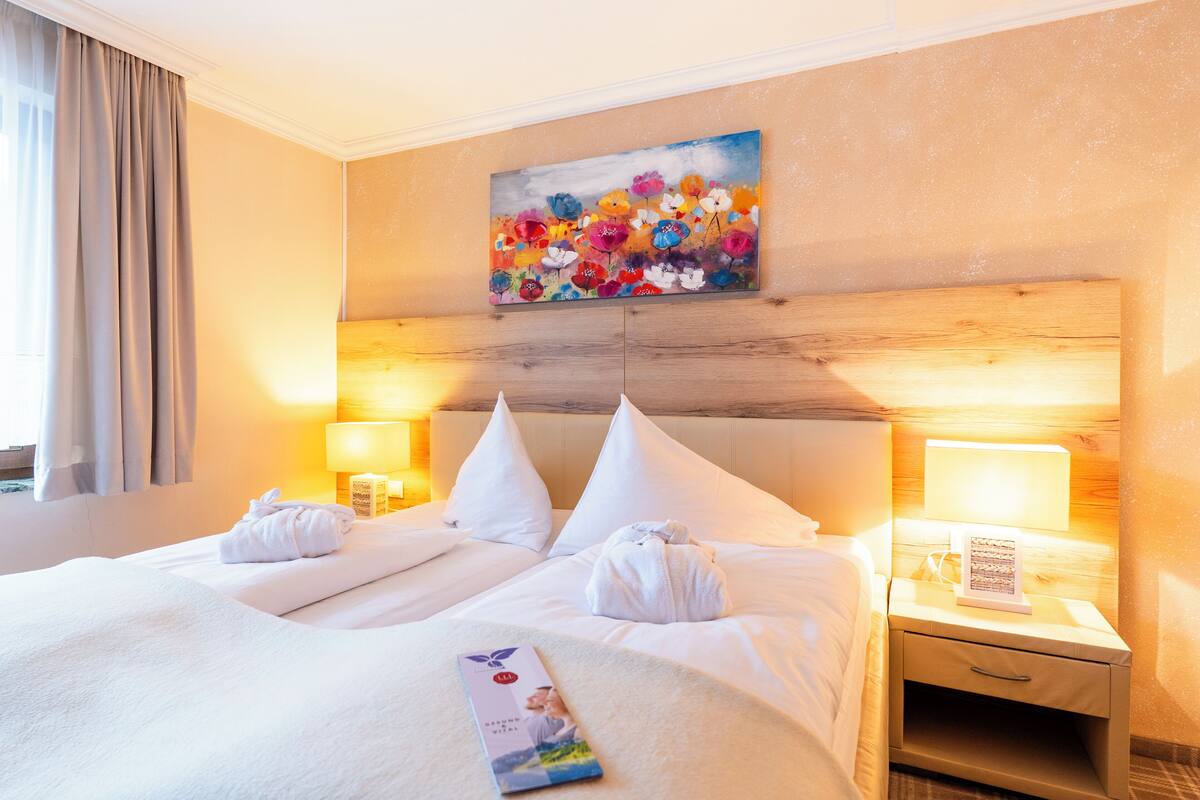 A hotel room with a large bed, two bedside tables with lamps, a painting on the wall, and a window with curtains.