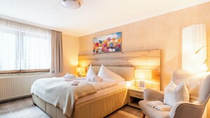 A hotel room with a large bed, two bedside tables with lamps, a chair, a painting on the wall, and a window with curtains. - DAS Hubertus Apartments in Schladming (Schladming)
