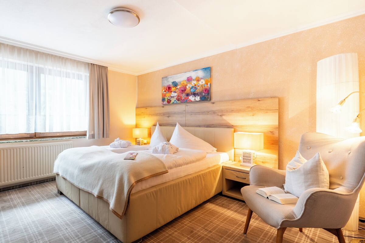 A hotel room with a large bed, two bedside tables with lamps, a chair, a painting on the wall, and a window with curtains.