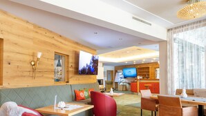 A modern hotel lobby with a wooden wall, a television, seating areas, and a dining space. - DAS Hubertus Apartments in Schladming (Schladming)