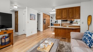 A modern living room with a flat-screen TV, wooden flooring, a wooden coffee table with decorative items, a beige sofa with a patterned pillow, and a kitchen area with wooden cabinets and a central island.