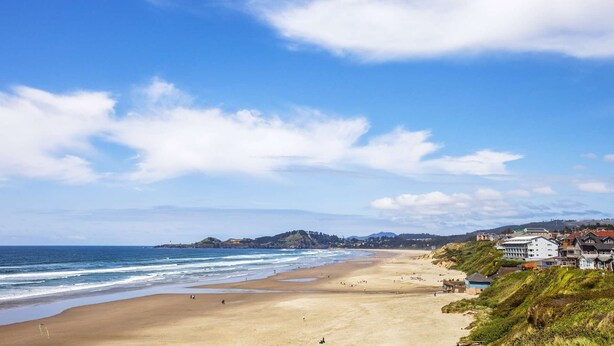 A coastal beach scene with a sandy shore, a few buildings, and a lighthouse in the distance.