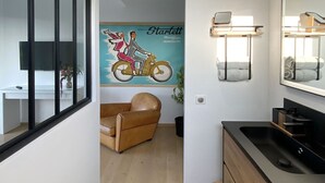 Une salle de bain moderne avec un lavabo noir, un meuble-lavabo en bois et un grand miroir. Une chaise beige se trouve dans la pièce adjacente, et une affiche de Starlett est accrochée au mur.