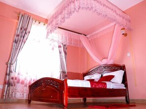 A bedroom with a four-poster canopy bed, a wooden headboard, and a red accent pillow. - Hotel Lintons Busia (Busia)