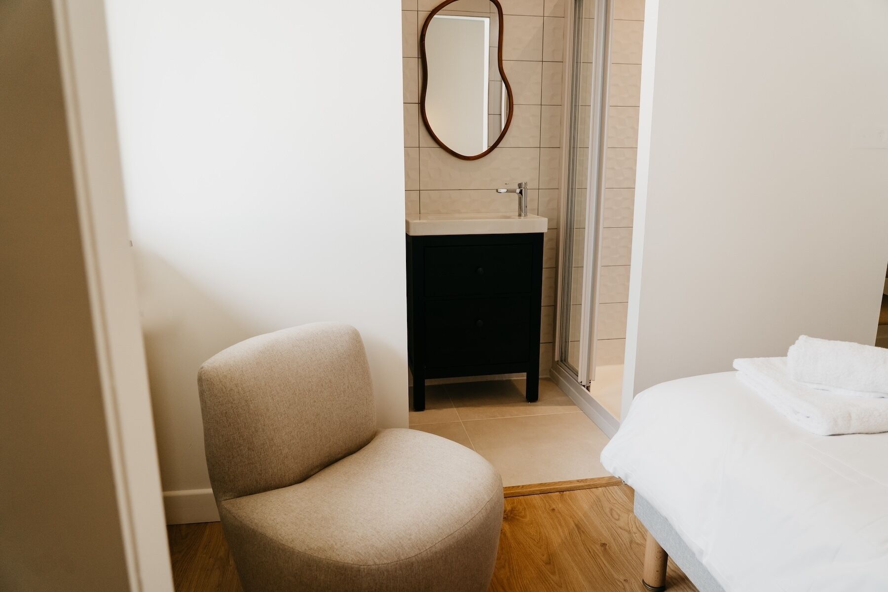 Un dormitorio con cama, una silla, una mesita de noche pequeña, un espejo y zona de ducha.