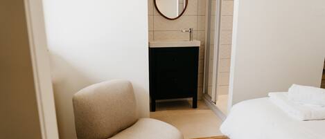 Un dormitorio con cama, una silla, una mesita de noche pequeña, un espejo y zona de ducha.