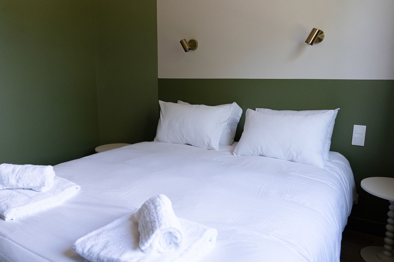 A neatly made bed with white linens and pillows, a bedside table with a lamp, and a wall-mounted light fixture.