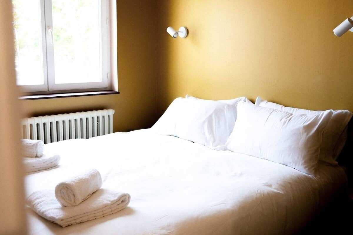 A neatly made bed with white linens and rolled towels, a window with blinds, and a radiator under the window.