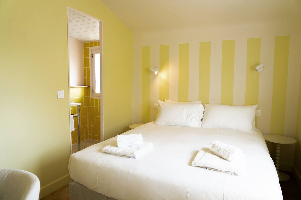 A hotel room with a large bed, two pillows, and two towels. There is a small bedside table and a chair. The walls are painted yellow and white.