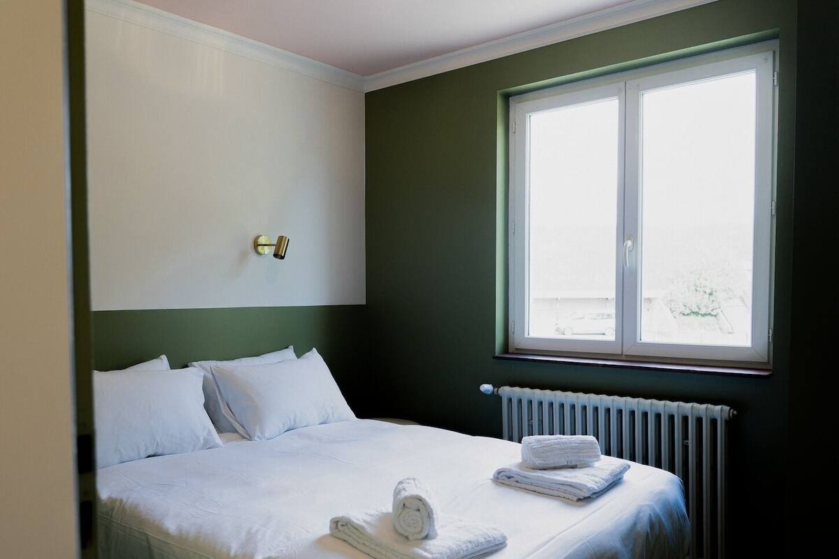 A hotel room with a bed, white linens, a window with a view, a radiator, and a wall-mounted light fixture.